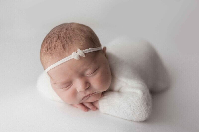 Close-up of a newborn baby girl sleeping peacefully on her tummy, wearing a white knit outfit and a delicate headband with a small bow, set against a soft, white background.
