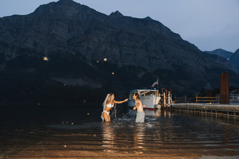 Bride in a bikini with veil and groom splashing water at each other at their destination wedding.