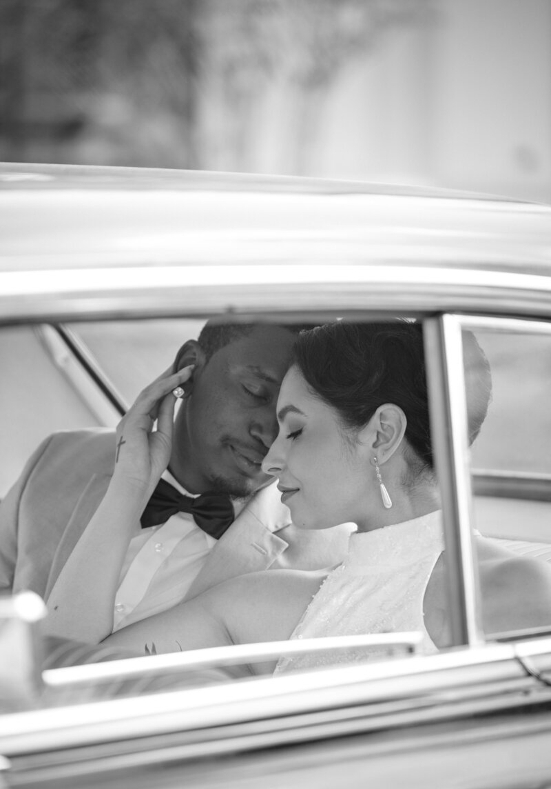 A couple sits in a vintage car on their wedding day and snuggles close