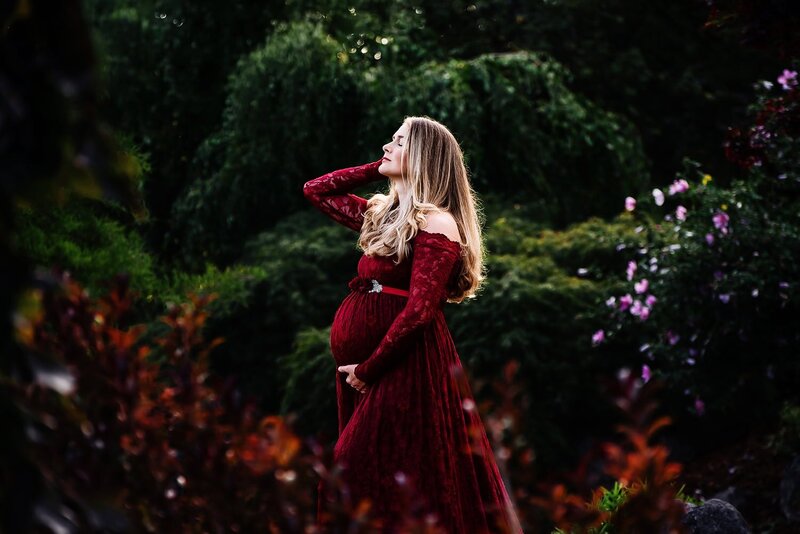 Expecting mother in a red lace dress striking a dramatic pose during an outdoor maternity session in the garden at Deer Lake Park.
