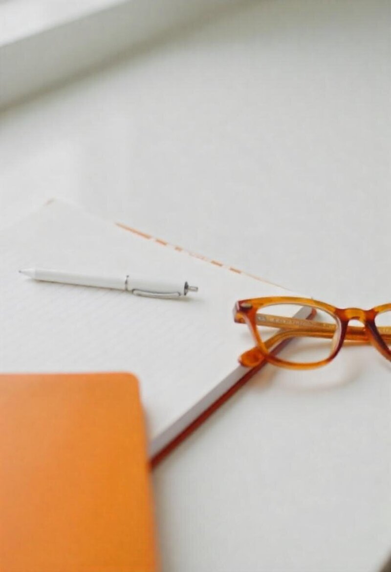 Notebook with a white pen and amber glasses on a white surface, with an orange journal partially in frame.
