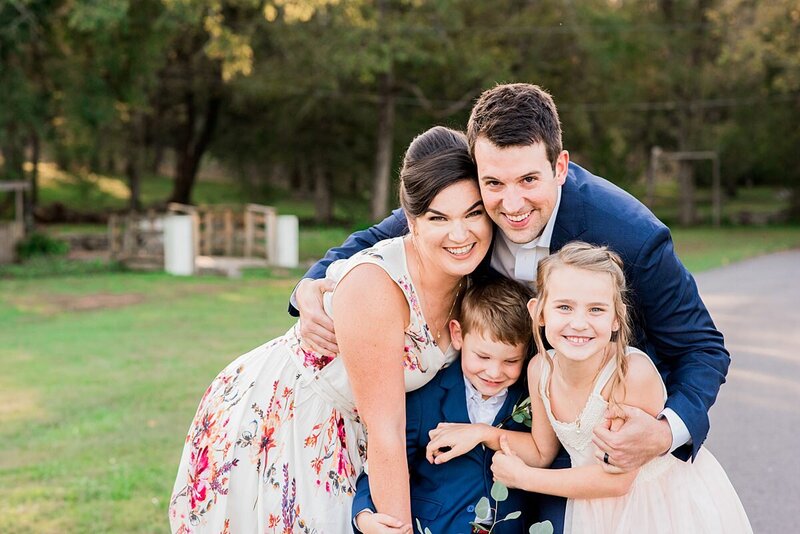 The bride and groom huddle together with the flower girl and ring bearer and they all smile at the camera. The bride is wearing a sleeveless white dress with an pink and brown floral pattern. The flower girl is wearing a white sleeveless sundress. The ring bearer and groom wear matching cobalt suits.
