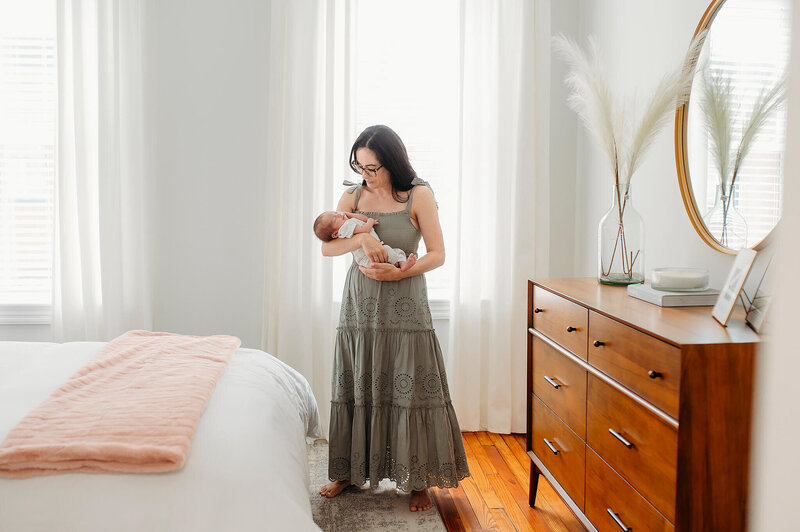 Woman in white sweater sitting on bed holding newborn baby rubbing noses with blonde girl in purple dress in Baltimore Maryland