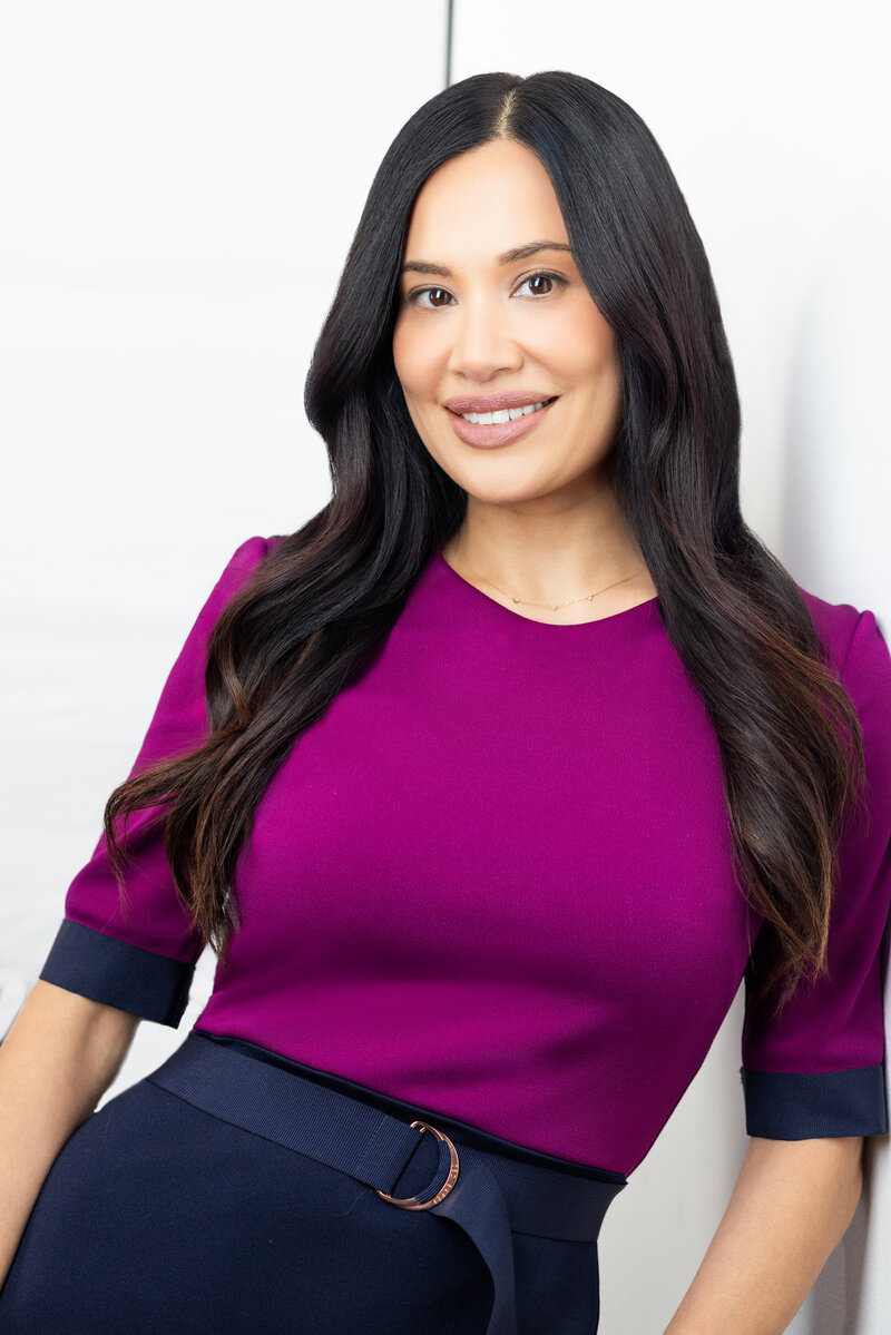 Woman in business attire with long black hair leans up against a white wall and looks at the camera during a business headshot session with philippe studio pro.