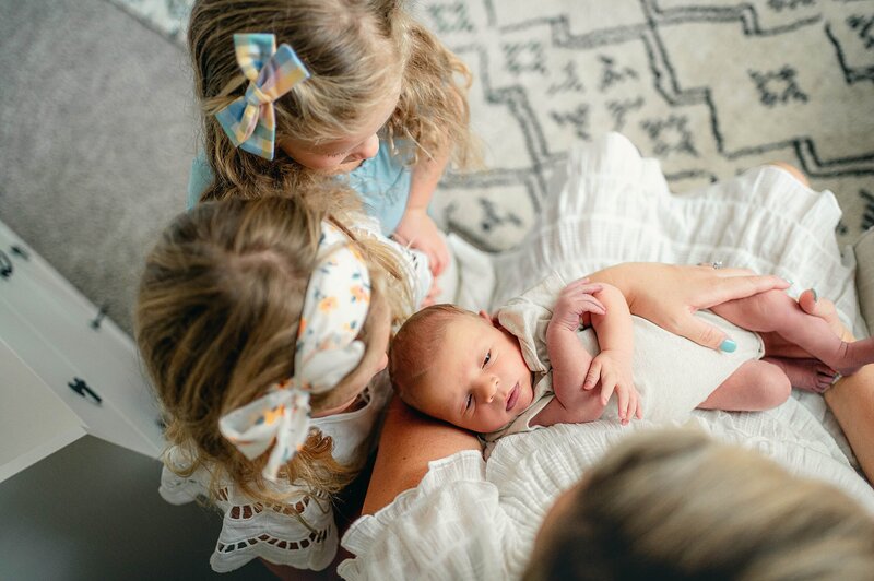 mom sitting and holding baby while big sisters watch at their Houston Newborn Photography session