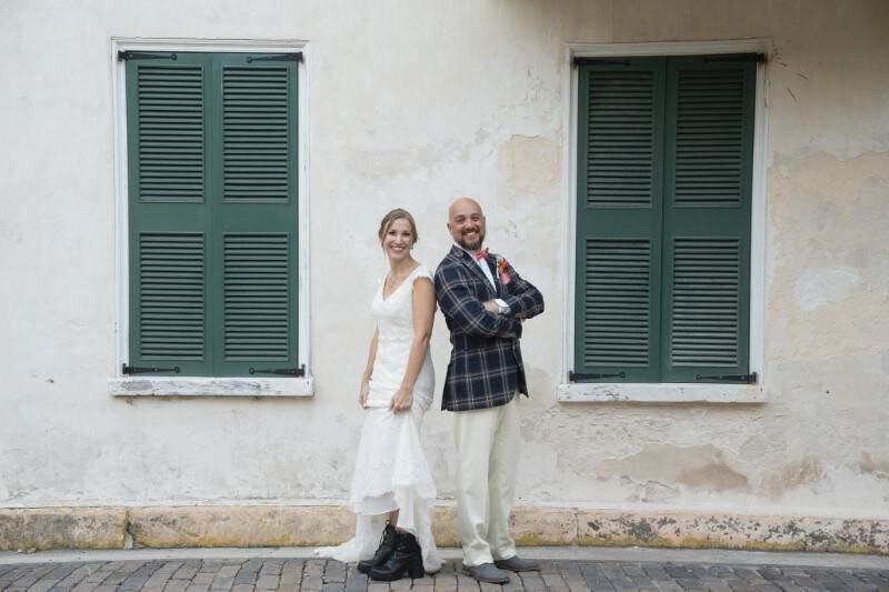 A bride in a white dress and boots and a groom in a jacket and slacks smile and pose back to back in front of a wall with green shutters