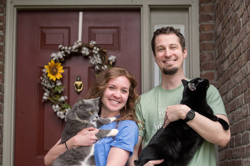 Couple holds 2 cats standing  outside red front door