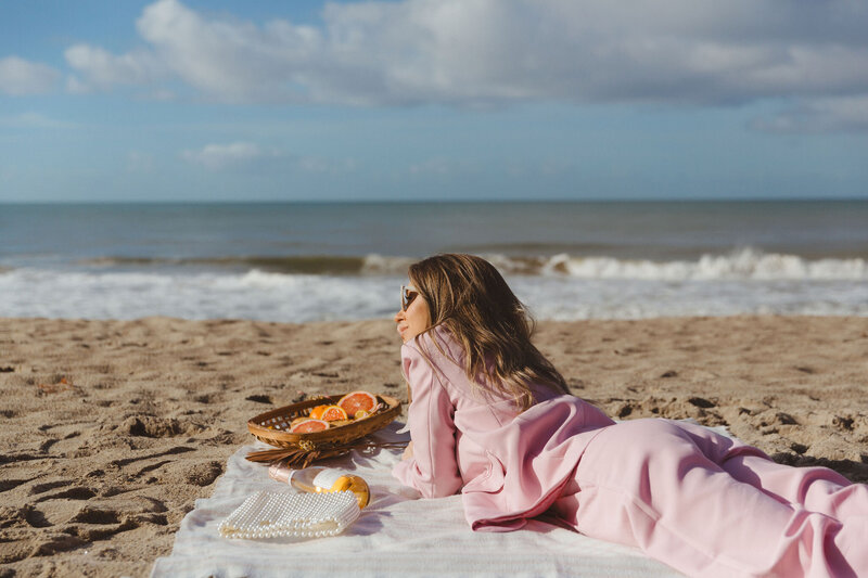 Jessa Jones, the owner of Shindig Social, lying on a beach in San Clemente, California. She is wearing a pink outfit and sunglasses, enjoying a picnic with fresh grapefruit and a pearl handbag on a blanket. The ocean waves and blue sky create a serene backdrop, highlighting Shindig Social's emphasis on authentic gatherings.