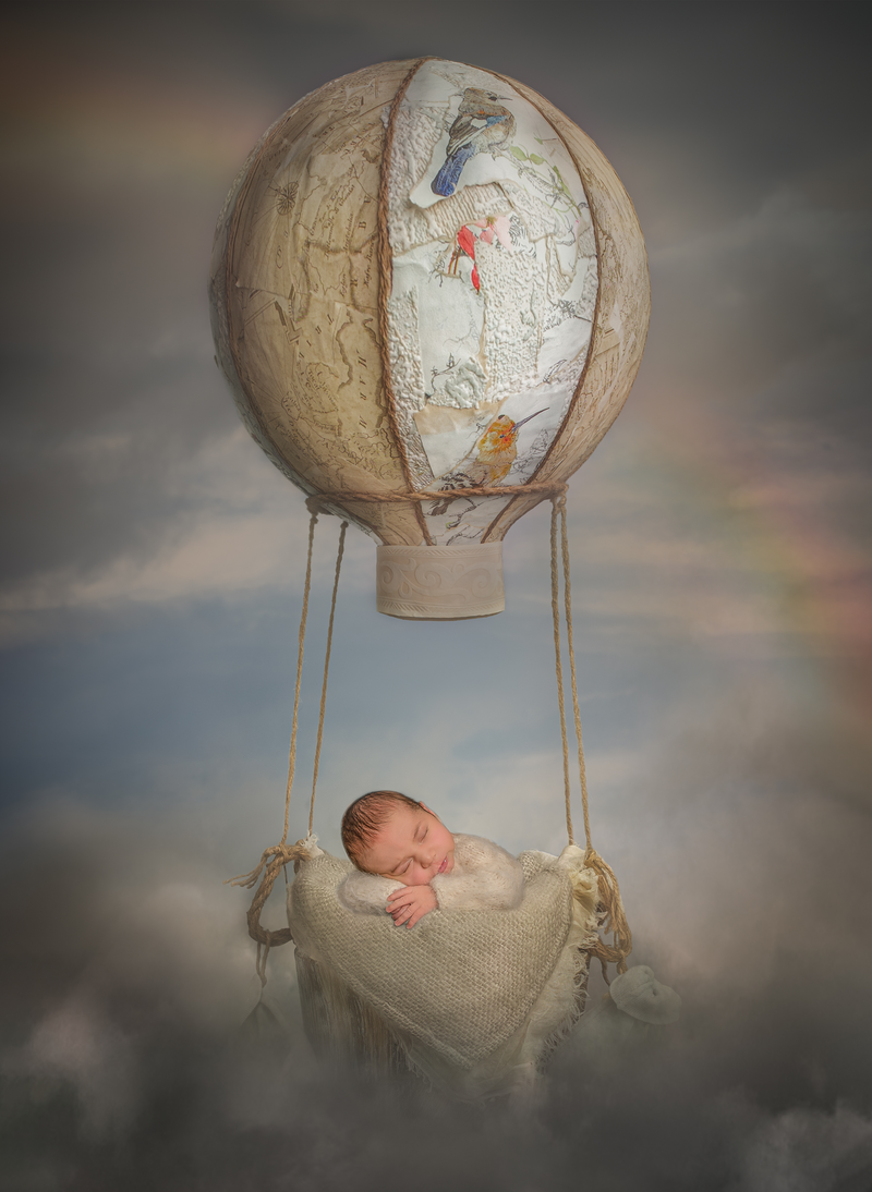  "Newborn baby peacefully sleeping in a miniature hot air balloon prop, surrounded by soft clouds, captured in a whimsical fine art photography session in Ottawa."