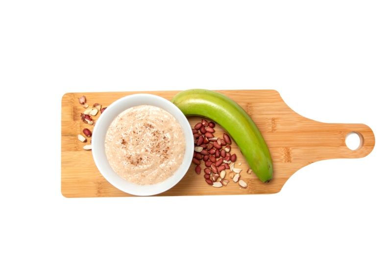 Bowl of peanut plantain porridge on a rectangular wooden board, with peanuts, and a green plantain.