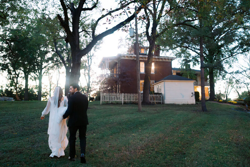 Newlyweds walk hand-in-hand towards historic house at sunset