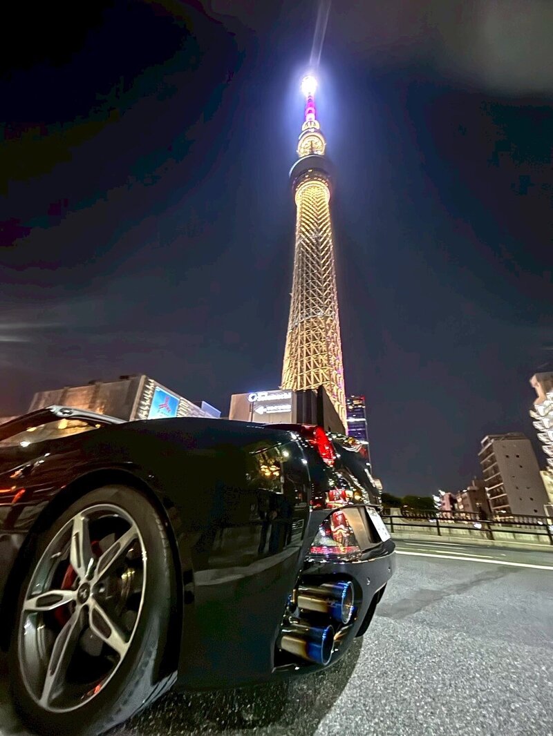 A glossy black Ferrari parked under the glowing Tokyo Skytree at night, reflecting city lights with a bold urban energy.