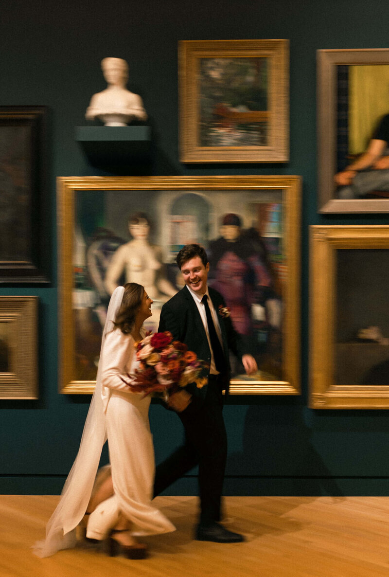 Bride and groom walking through an art gallery, with the bride holding a bold bouquet of rust, burgundy, and peach flowers. Editorial floral design by Springvale Floral, luxury wedding florist serving DC, Virginia, and Maryland.

