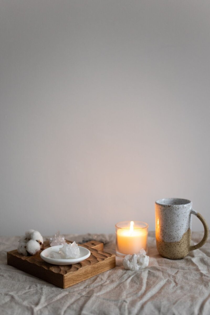 Minimalist wellness scene with a candle, ceramic mug, cotton sprigs, and white crystals on a wooden tray and linen cloth