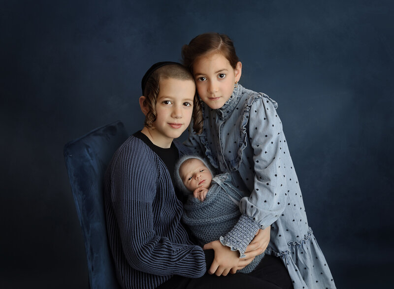 Brooklyn newborn photoshoot. Big sister and big brother are holding their baby brother. All are looking at the camera. Blue clothing and backdrop. Captured by best Brooklyn, NY newborn photographer Chaya Bornstein.