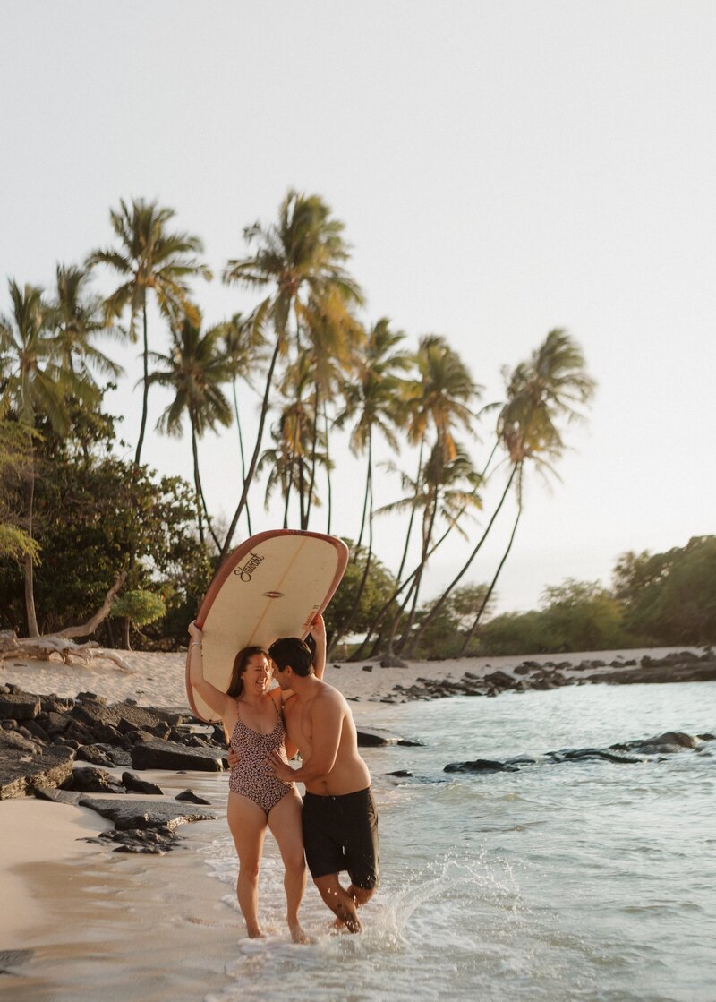 couple laughing and walking on beach holding a surfboard