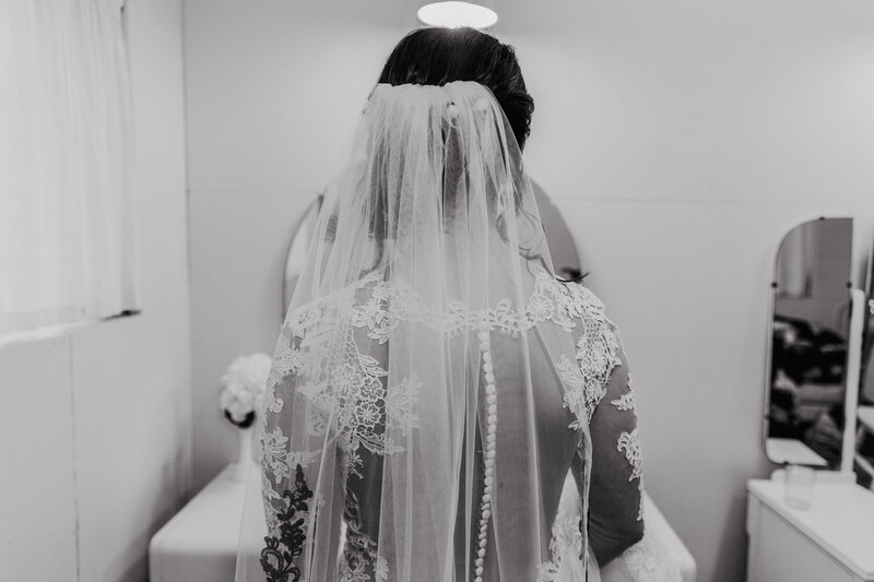 View of the brides lace button up gown and veil, as she looks at herself in the mirror 