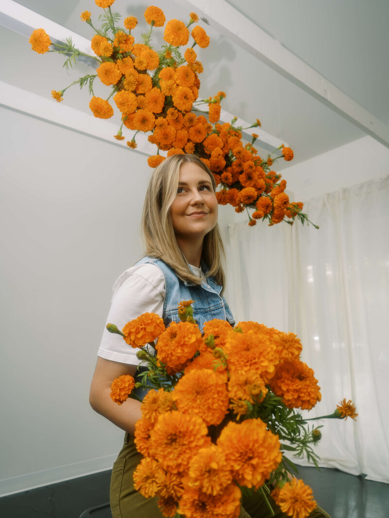 Eugene, Oregon florist, Hope, holds a bouquet and sits under a suspended floral arrangements. The flowers are all orange marigolds.
