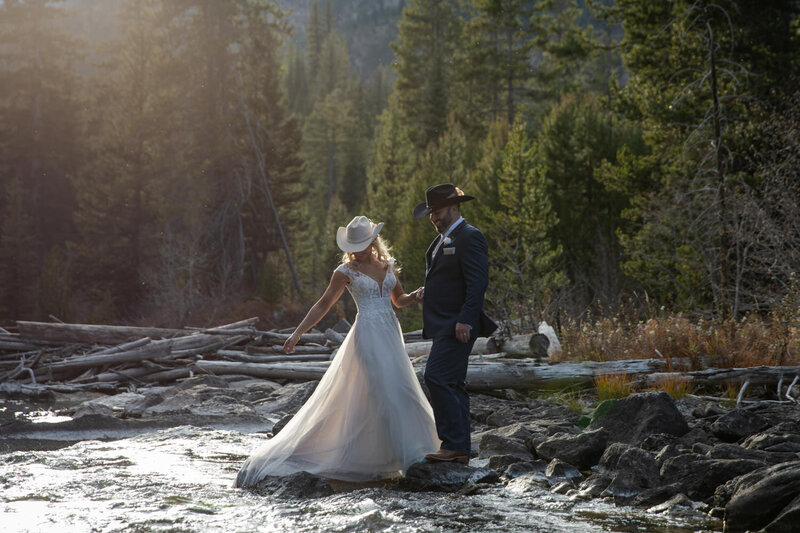 Bride and groom in cowboy hats crossing a rocky mountain stream, embracing a rustic and adventurous elopement in nature.