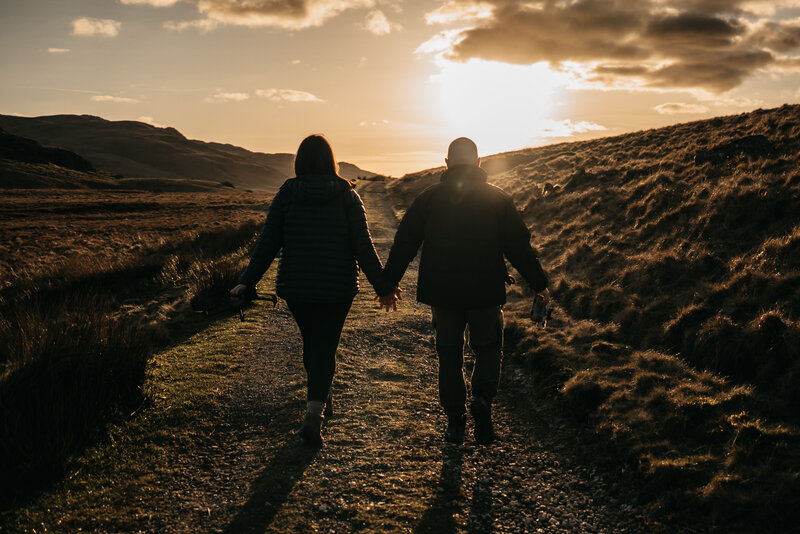 A couple walks hand-in-hand down a sunlit path through the Lake District hills, silhouetted by golden hour light. Captured by a wedding photographer and videographer duo specialising in natural, storytelling imagery across the Lake District and destination weddings worldwide.