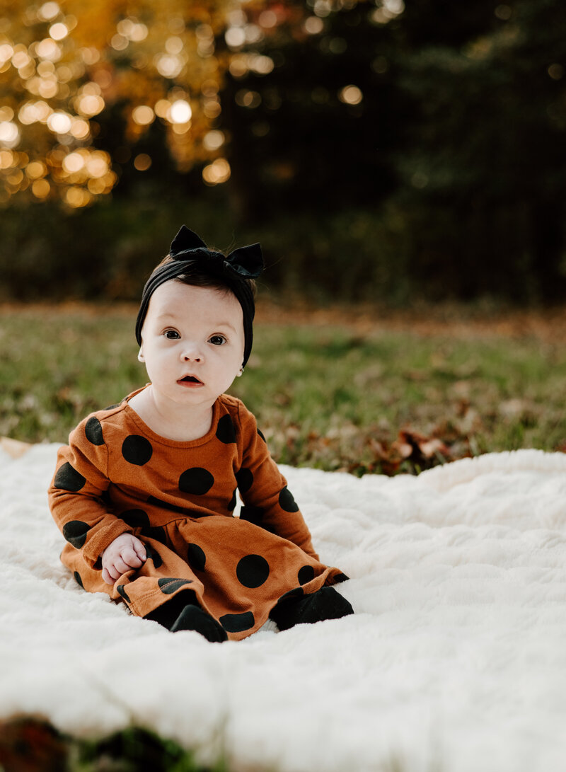 6 month old baby girl in orange and black polka dot dress sitting on white blanket in Annapolis Maryland park photographed by Bethany Simms Photography