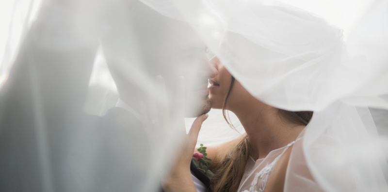 In a tender moment captured in a photograph, the bride and groom share a passionate kiss under a billowing veil. The veil, sheer and ethereal, envelops them both in a soft embrace, creating a sense of intimacy and romance. Their faces are illuminated by the gentle light filtering through the veil, highlighting the joy and love radiating from their expressions. With their eyes closed and lips locked in a tender embrace, they are lost in the magic of the moment. The scene evokes a timeless sense of romance and captures the profound connection between the bride and groom on their wedding day.