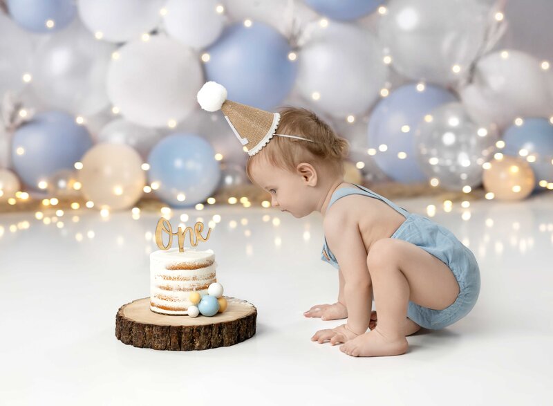 Baby boy looking at his birthday cake with white, tan and blue balloons in the background. This was a custom set decorated by Jenni Nikki at her Woodstock photo studio near Atlanta.