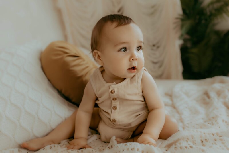 A baby in a beige outfit sits on a soft, textured blanket with a pillow and plants in the background.