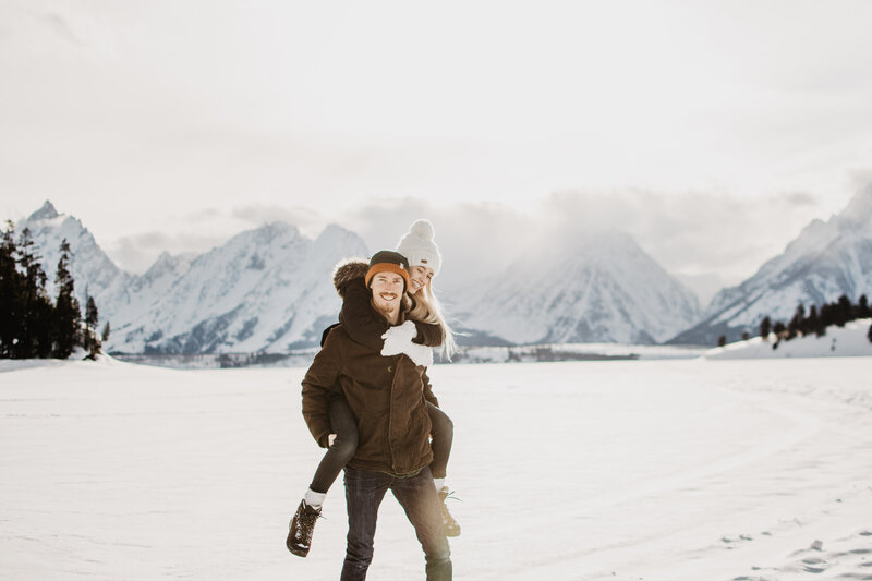 winter engagement session with jackson hole photographers capturing couple piggy backing on jackson lake on a sunny winter day