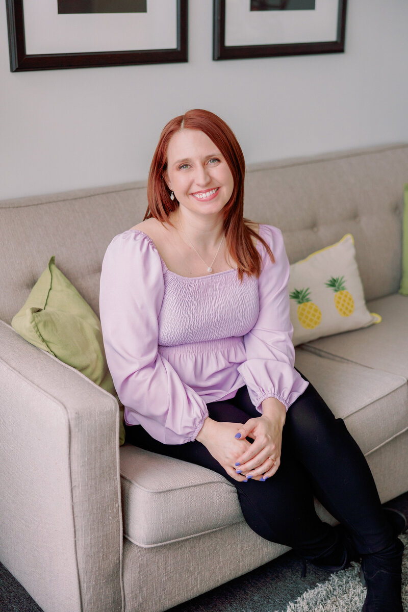 A person poses on a couch while wearing a purple shirt.