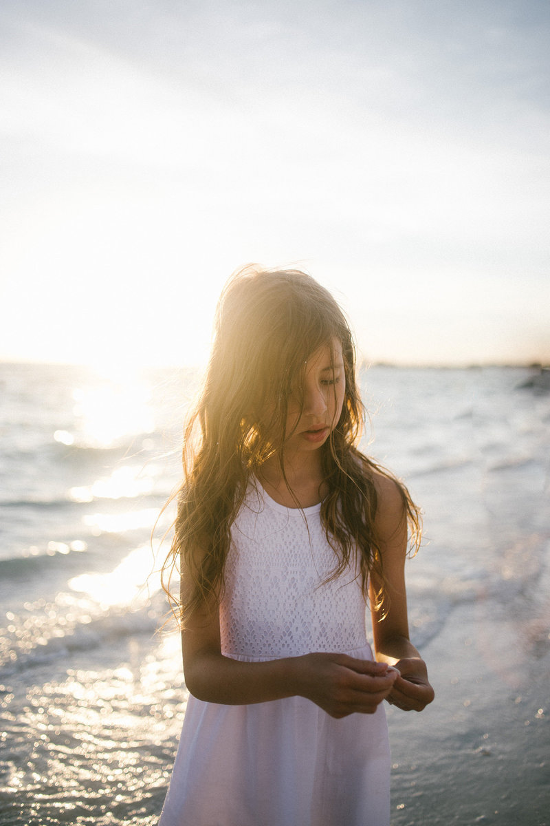 Little girl holding a sea shell during sunset at a beach wearing a white dress