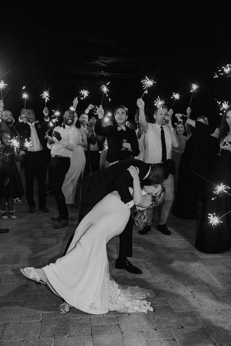 A couple kisses at their reception while their friends and family hold sparkers behind them.