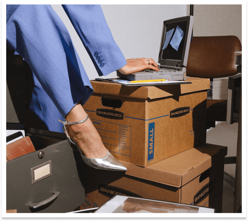 A young woman's foot in a silver heel resting on filing boxes with her old laptop with sticky notes everywhere.