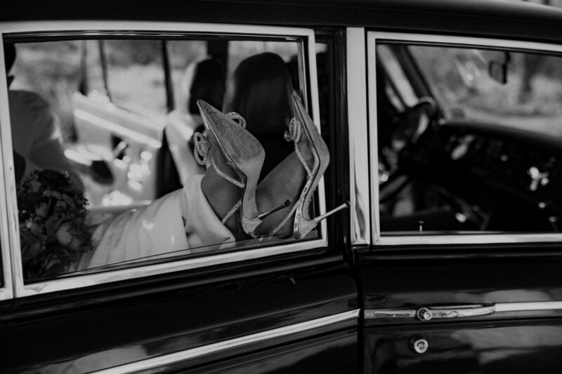 Bride's glamorous high heels peeking out of a classic car window during stylish elopement exit in black and white photo.