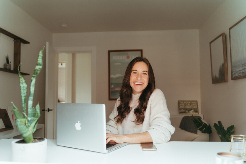Woman with brown curled hair wearing a beige sweater sits at desk on her MacBook and smiles at the camera