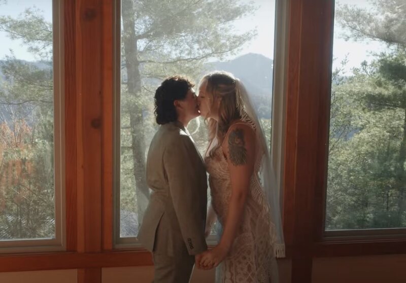 A newlywed couple kissing in front of windows overlooking the mountains 