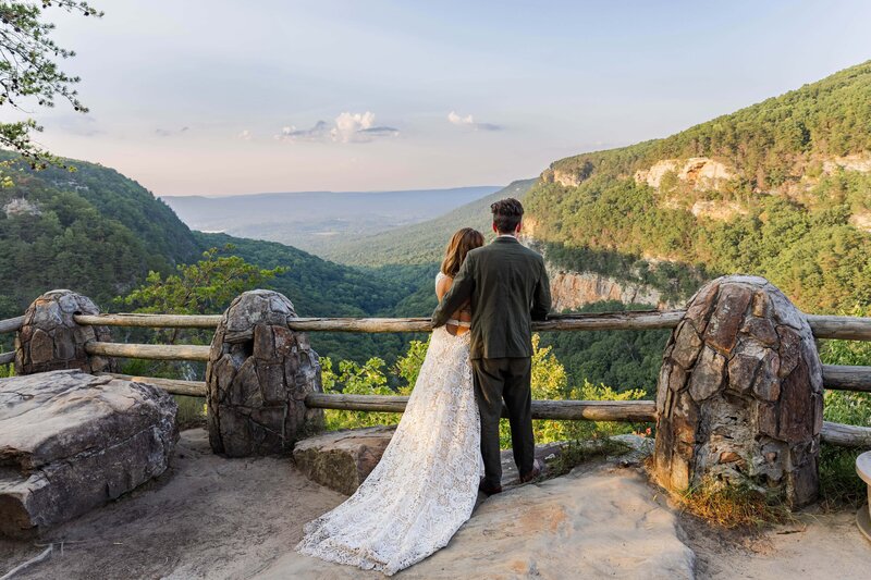 A bride and groom stand at an overlook looking out enjoying the views. Their backs are facing the camera with the grooms arm wrapped around the bride. The bride is wearing a lace white wedding gown with a train and the groom is in an olive green suit. There are rock and tree covered mountains in the distance with the sky turning pink from the sunset.