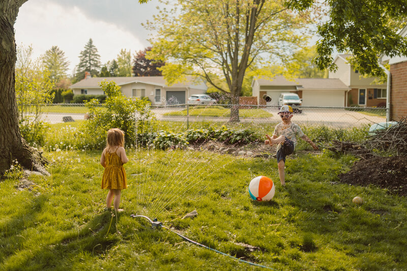 young brother and sister play in their backyard, kicking a beach ball back and forth over a sprinkler