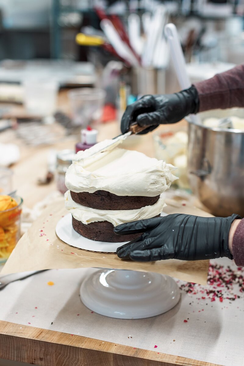 Baker frosting a two-layer gluten-free chocolate cake at Grain Artisan Bakery in Snohomish, Washington, highlighting their handcrafted cakes made with care and clean ingredients.