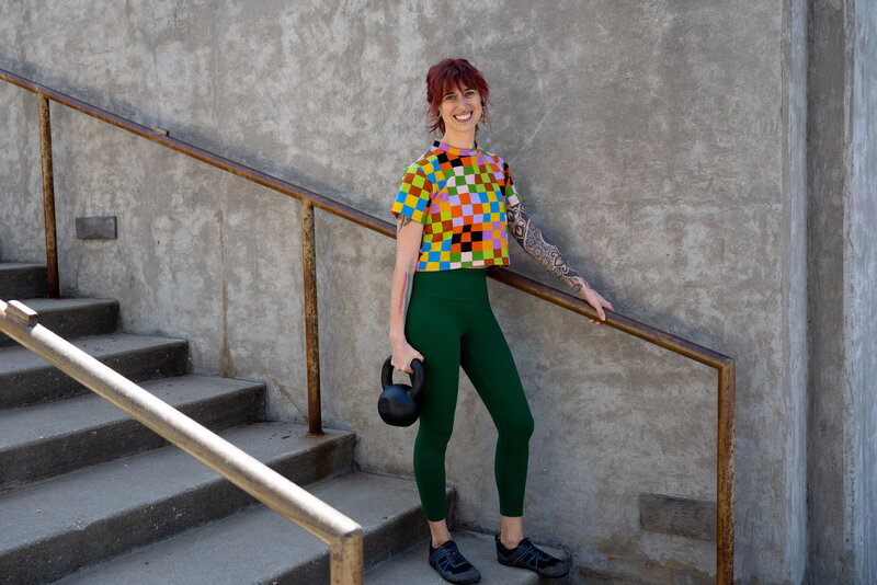 a woman in a rainbow checkered shirt hold a kettlebell while walking down steps to go to a strength training program