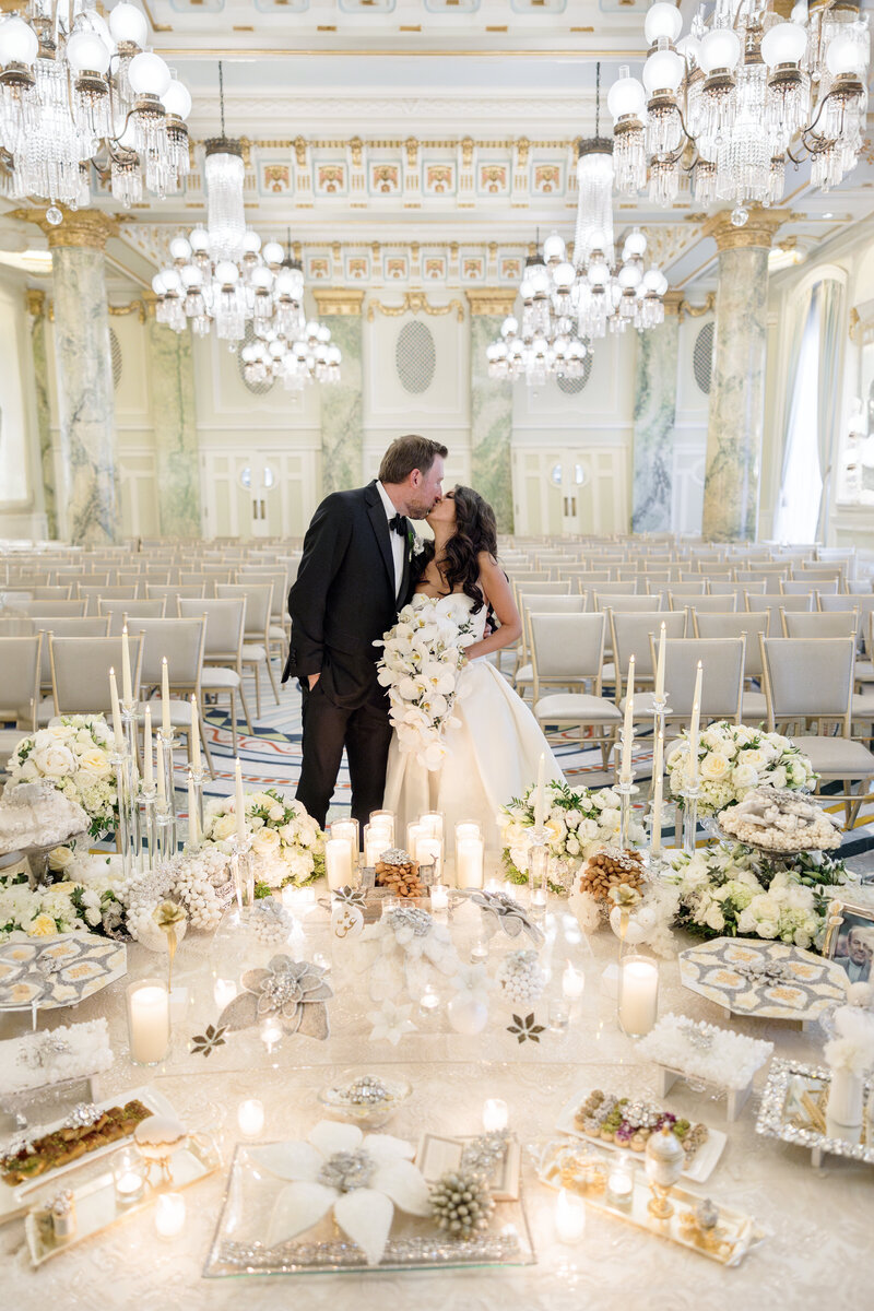 A bride and groom kiss in an ornate hall decorated for a wedding. They stand surrounded by flowers, candles, and elegant table settings. Rows of empty chairs fill the background, and chandeliers hang from the high ceiling.