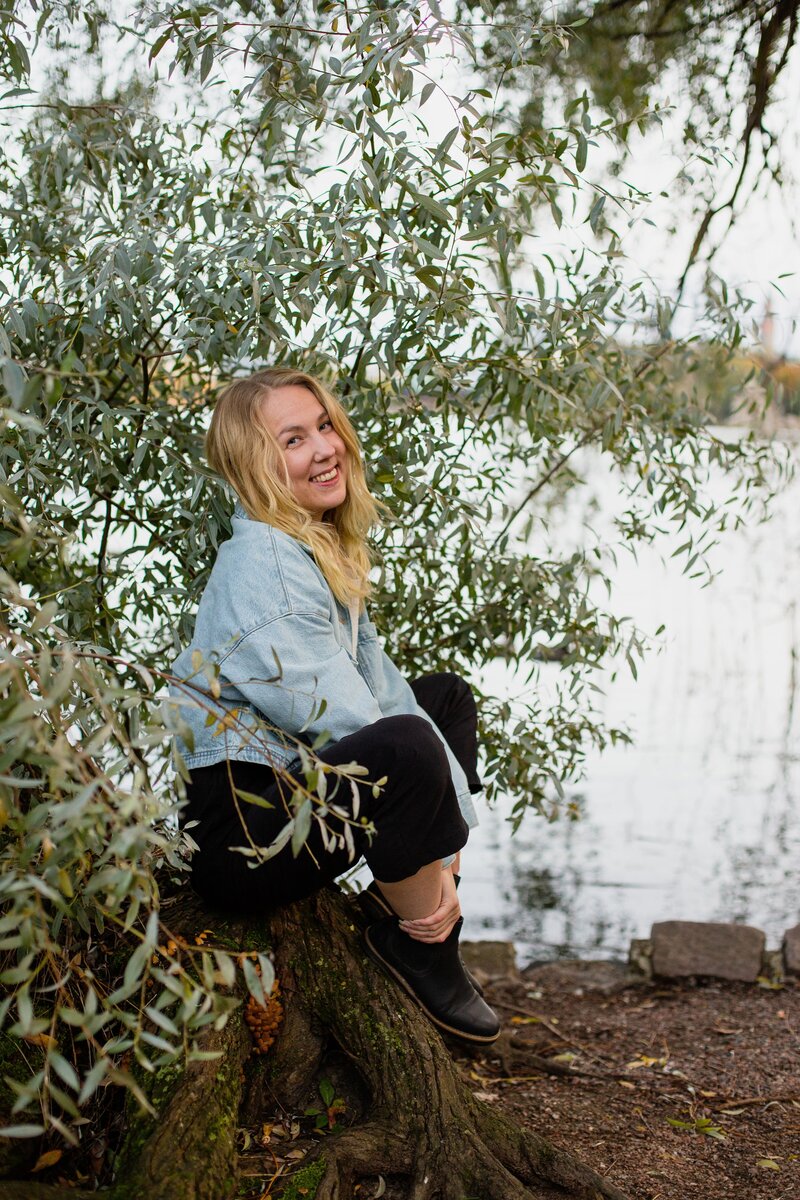 mielen paremminvoinnin ja kehomieliyhteyden valmentaja  istuu puun rungon päällä järvimaisema taustalla hymyillen