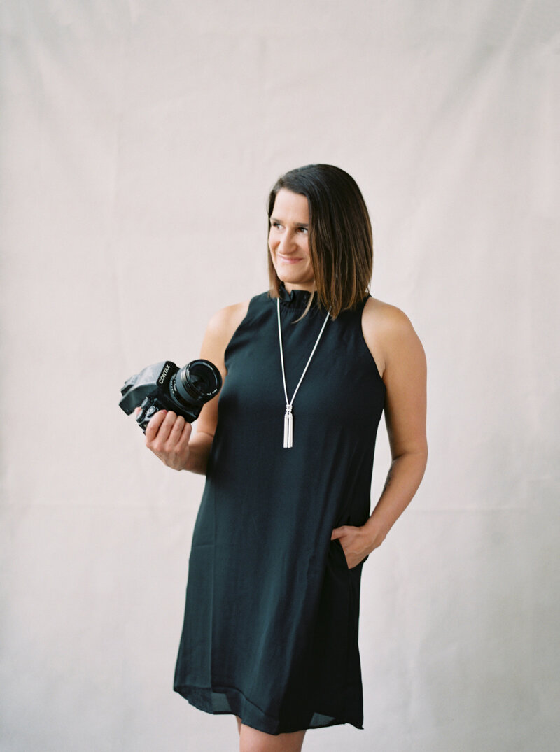 Courtney Bowlden smiling at camera in front of a white wall holding a camera in one hand and the other on her hip