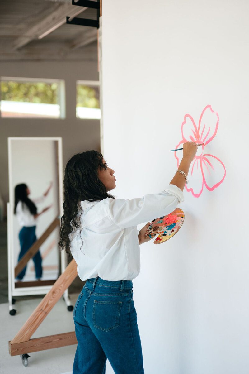 A woman in a white shirt and jeans paints a pink flower on a white wall, holding a palette. A mirror reflects her image, suggesting focus and creativity.