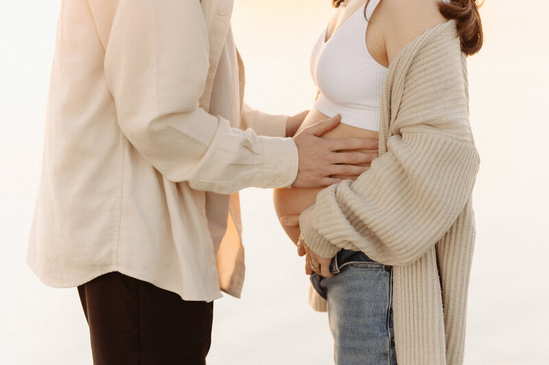 Close-up maternity moment of expecting parents at Coronado Beach, with the father’s hands on the mother’s belly during golden hour. Captured by San Diego Family Photographer Yuka Schad.