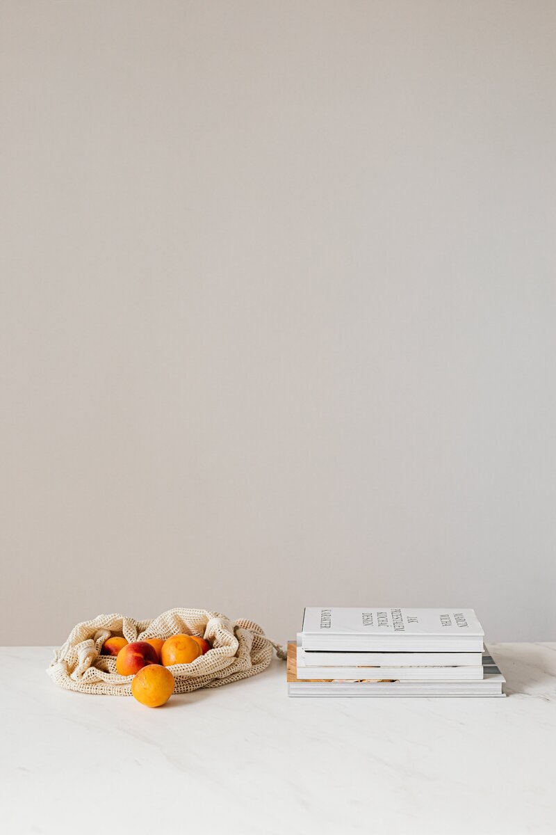 stack of four books, cloth produce bag opened with fruit, sat upon white countertop with beige backdrop