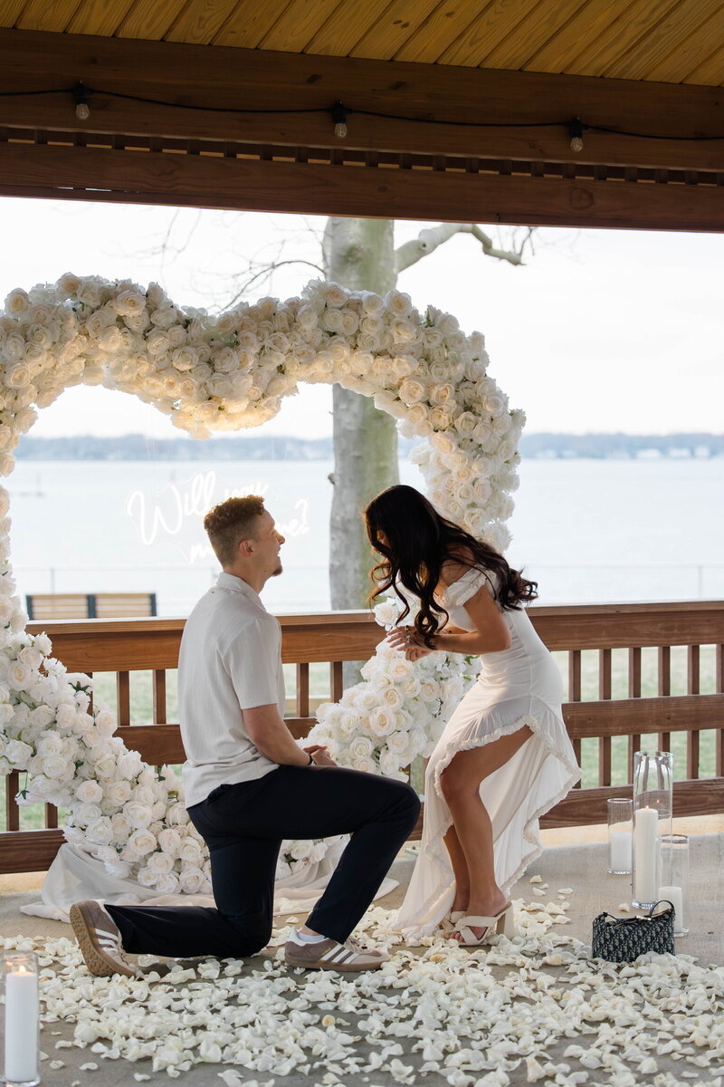 Romantic proposal by the water with a groom-to-be on one knee in front of a heart-shaped white flower arch, as his partner smiles in surprise. Styled by Yours Truly, a wedding planner and designer in New Jersey.
