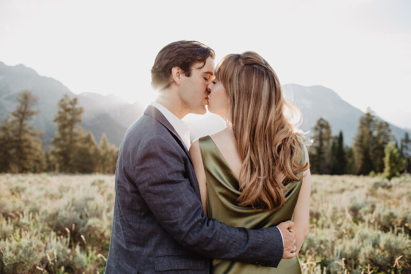 jackson hole wedding photographers captures engagement pictures with man and woman kissing at sunset with the sun shining over the mountains for their grand teton engagement pictures
