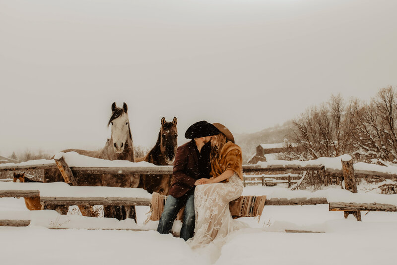bride and groom sitting on a wood fence with horses in the background while it is snowing during their colorado elopement