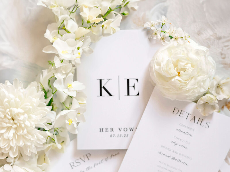 A subtle, elegant wedding invitation set with white flowers. The cards feature minimalist typography with initials and sections for vows, RSVP, and event details. The soft floral arrangement frames the stationery.